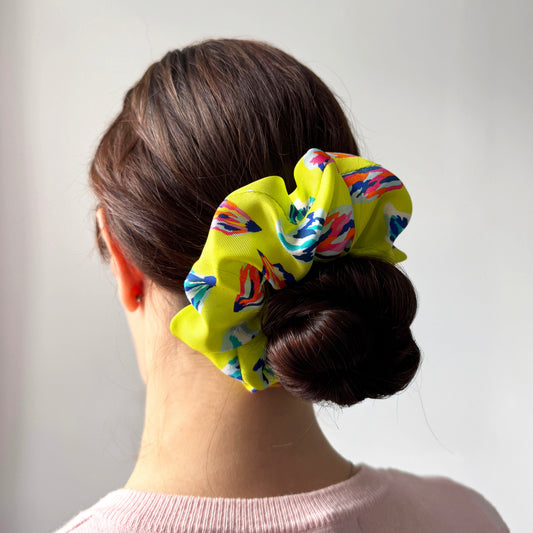 A woman shown from the back wearing a bright yellow fabric scrunchie with a colorful abstract pattern in red, blue, and white, styled in a low bun.