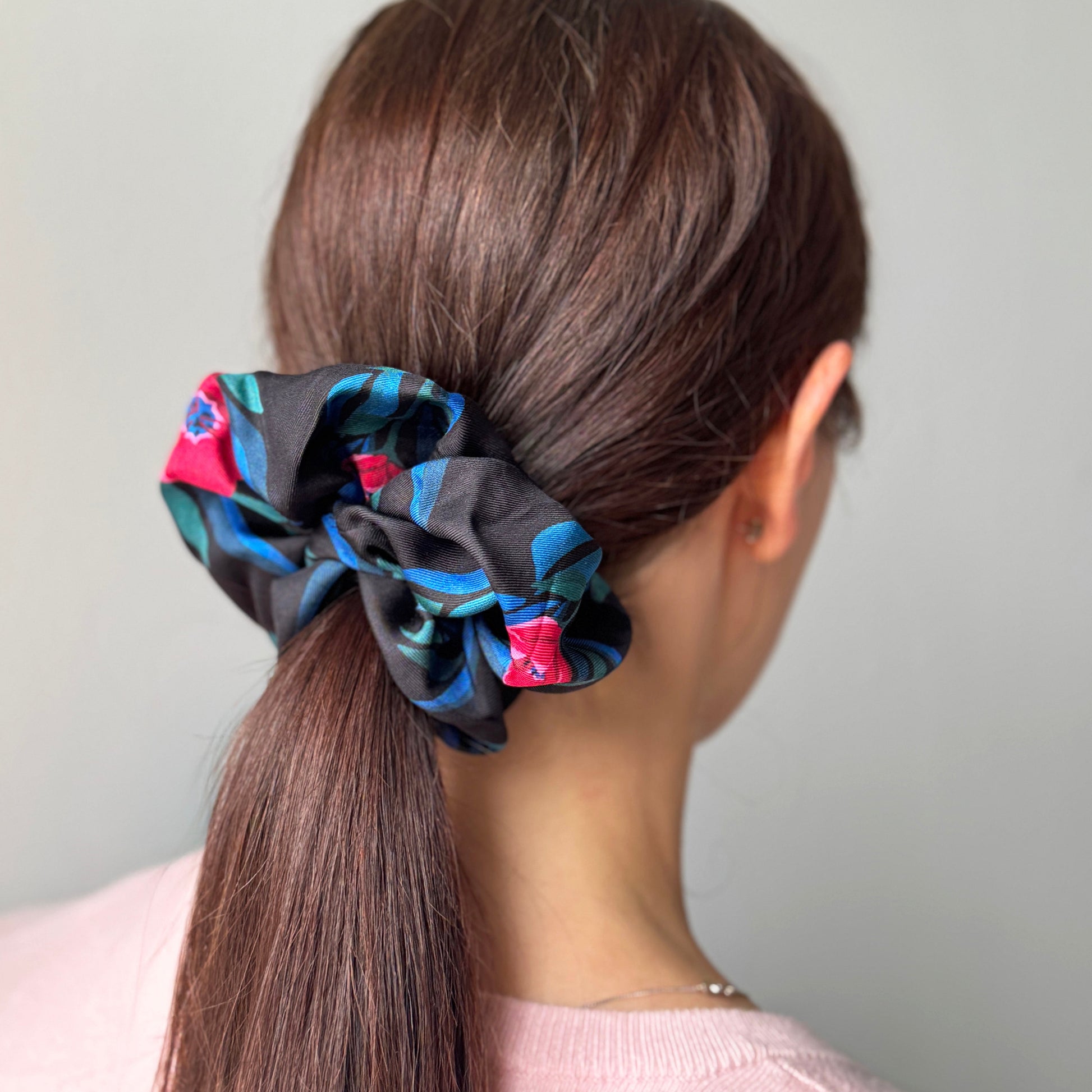 A black hair scrunchie with a blue, green, and pink pattern, shown on a model's ponytail from the back.
