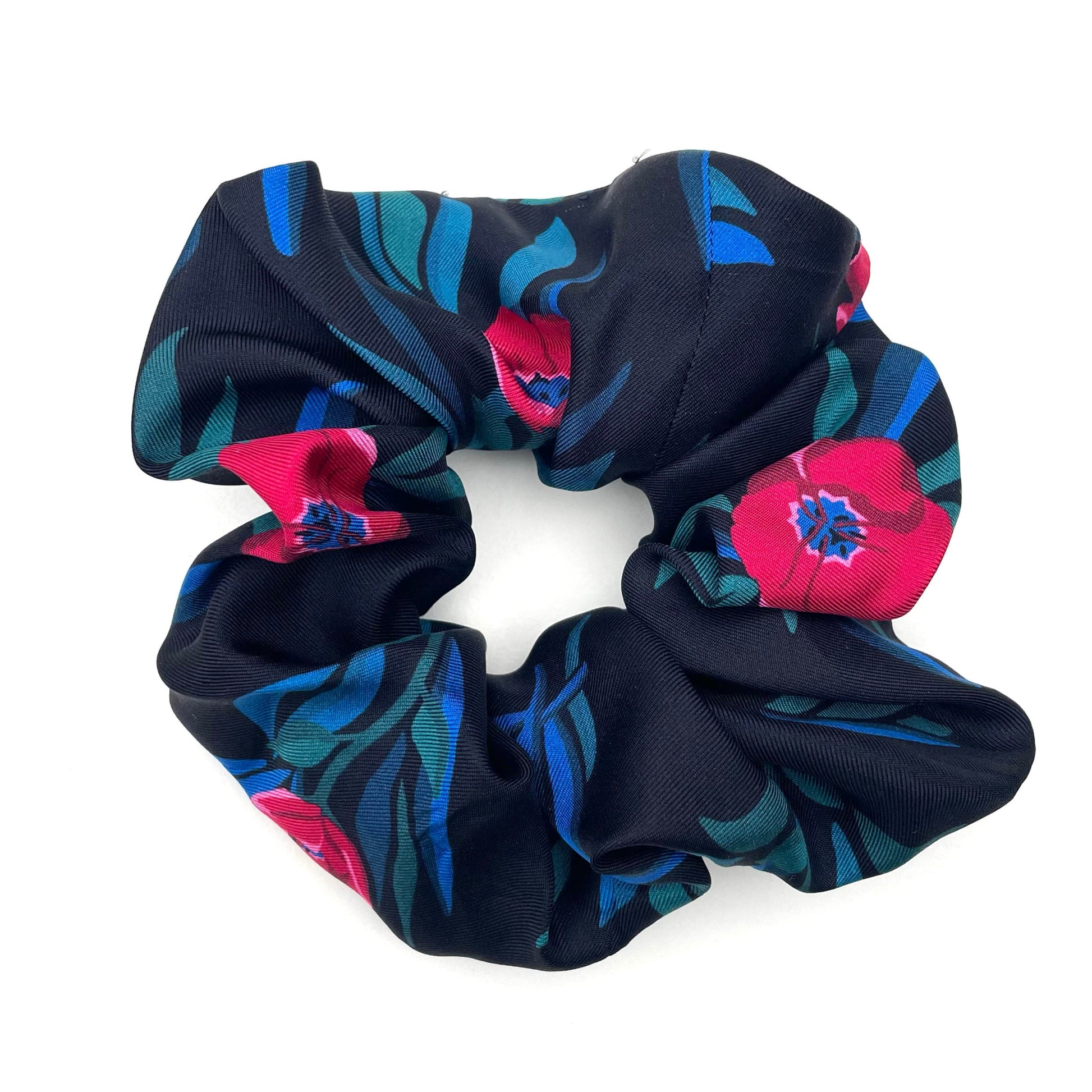 A fabric hair scrunchie with a black background and a bold floral pattern in bright pink and teal, photographed on a clean white background.