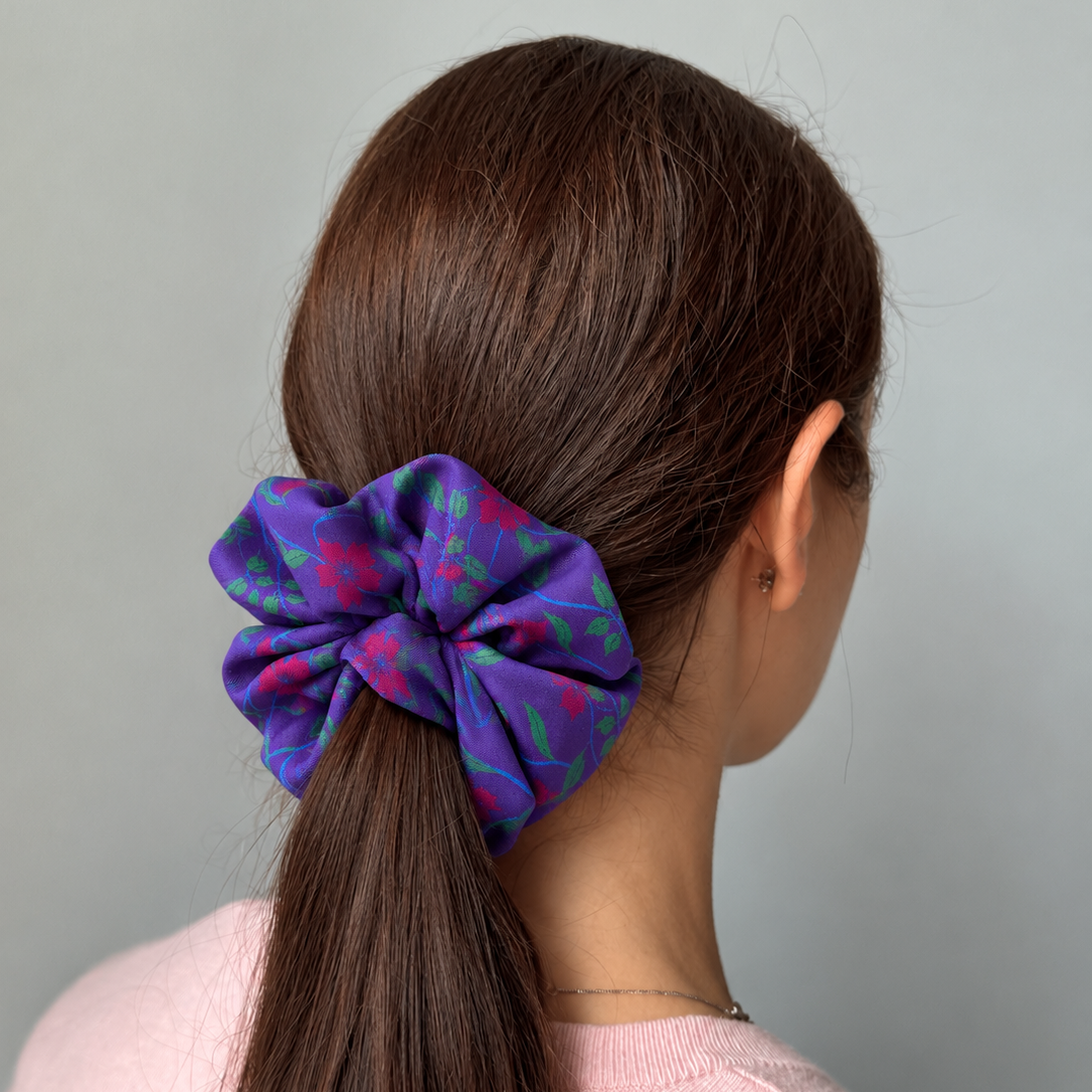 A purple hibiscus print hair scrunchie with a purple and pink floral pattern, shown on a model's ponytail from the back against a plain white background.