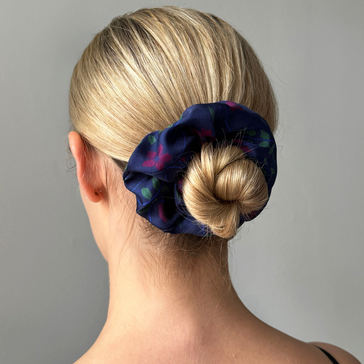 A blonde woman shown from the back wearing a dark navy blue silk scrunchie with a subtle pink and green floral pattern, holding her hair in a low bun.