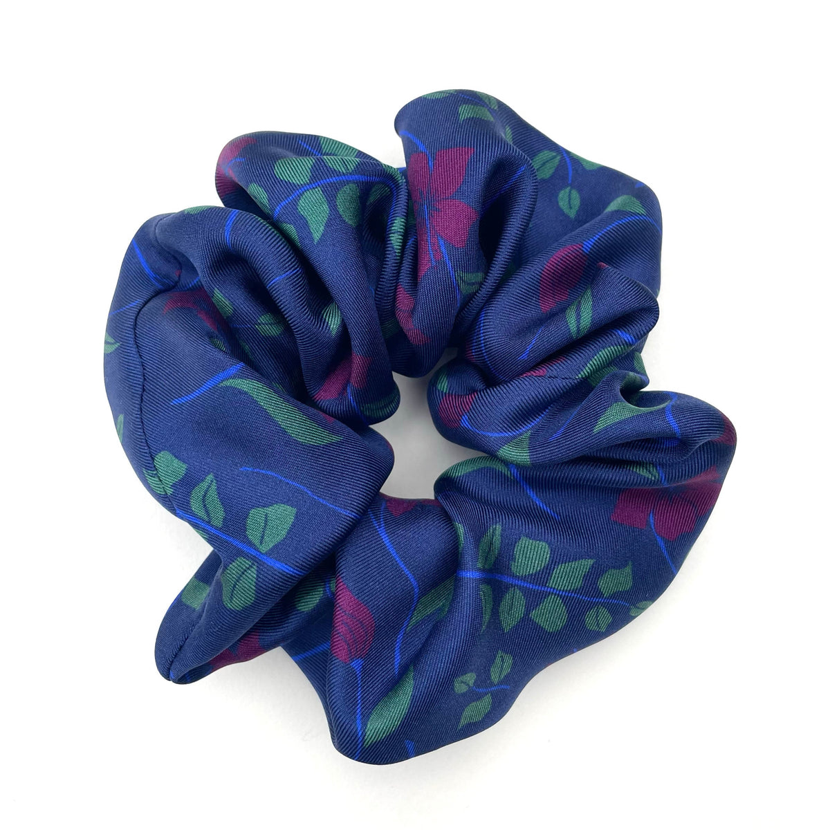 A silk hair scrunchie with a navy blue base, featuring a pattern of green leaves and purple flowers, photographed on a clean white background.