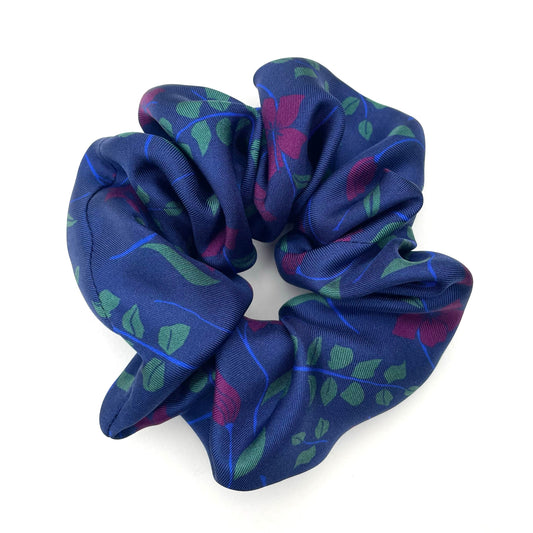 A silk hair scrunchie with a navy blue base, featuring a pattern of green leaves and purple flowers, photographed on a clean white background.