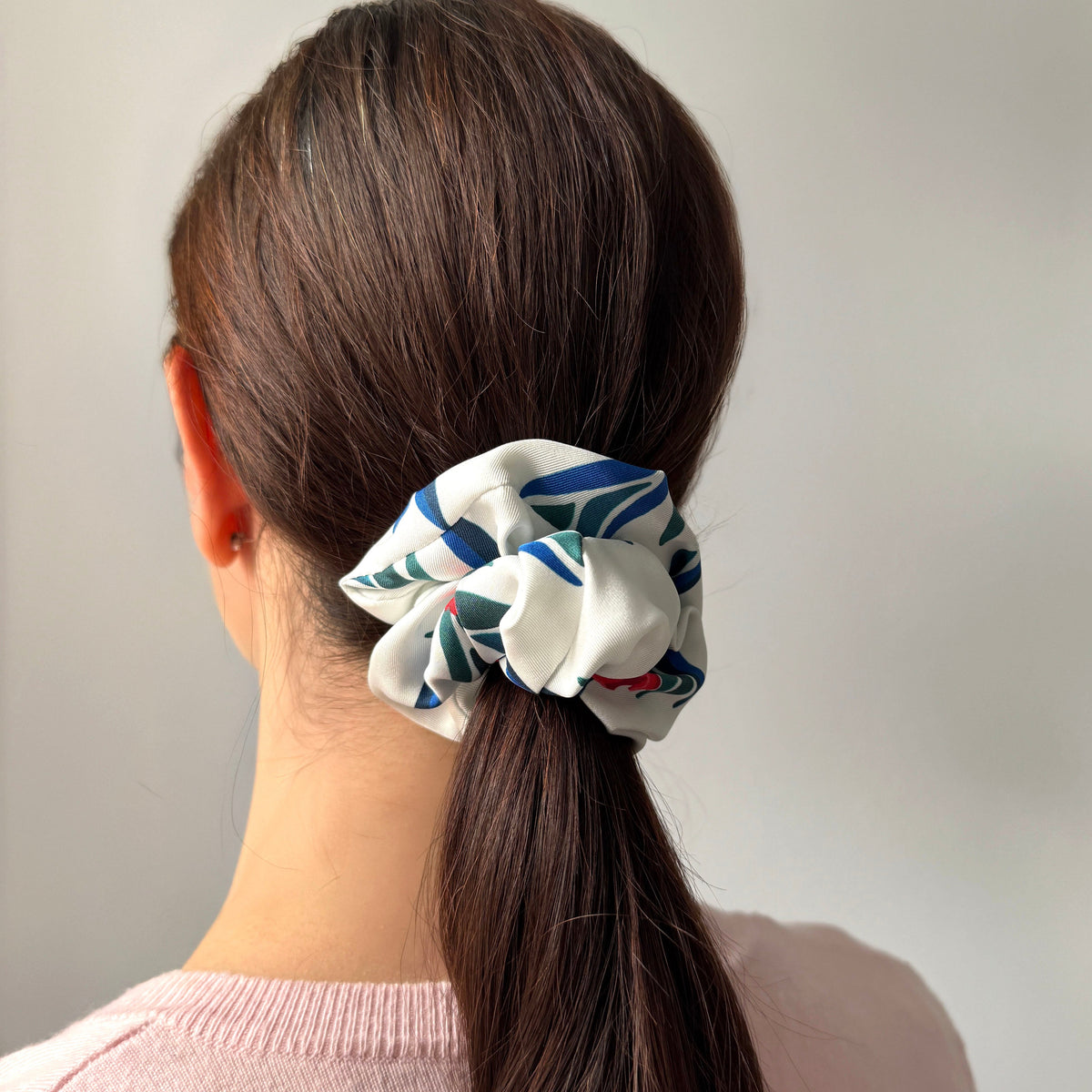 A woman viewed from behind wearing a white silk fabric scrunchie with a red, blue and green floral print  in her brown ponytail, paired with a light pink knit top against a plain light gray background.