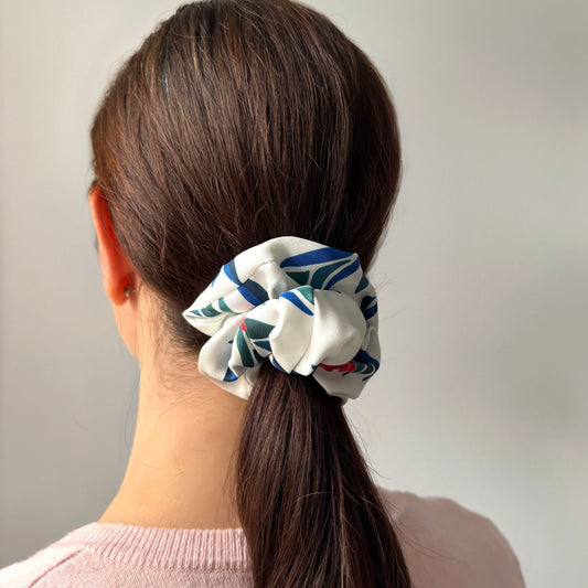 A woman viewed from behind wearing a white silk fabric scrunchie with a red, blue and green floral print  in her brown ponytail, paired with a light pink knit top against a plain light gray background.